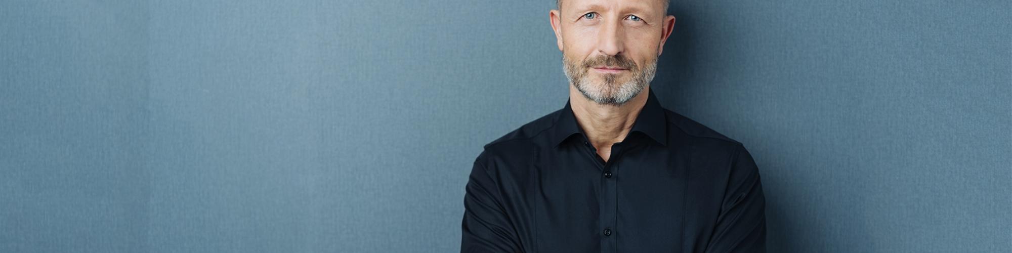 Vasektomie: Mann lächelt mit geschlossenen Lippen vor blau-grauem Hintergrund