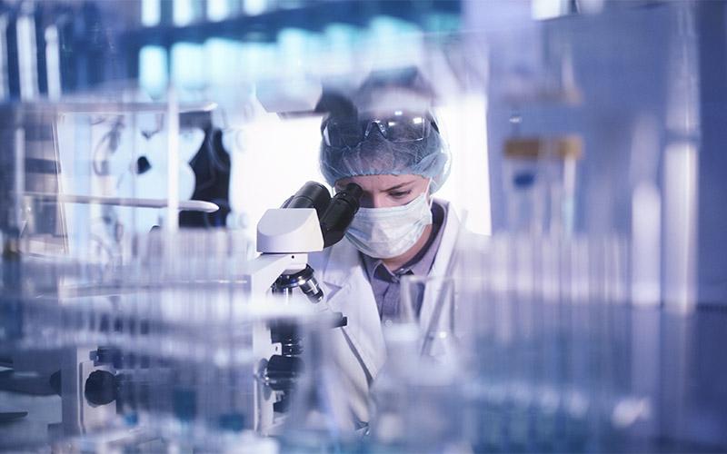 Mitarbeitender eines Labors mit Mundschutz und Haube hinter einer Reihe von Reagenzgläsern