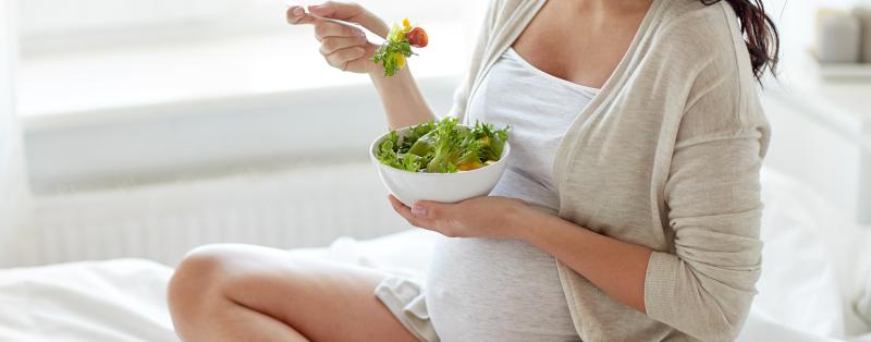 Schwangere Frau sitzt auf dem Bett und isst Salat aus einer Schüssel